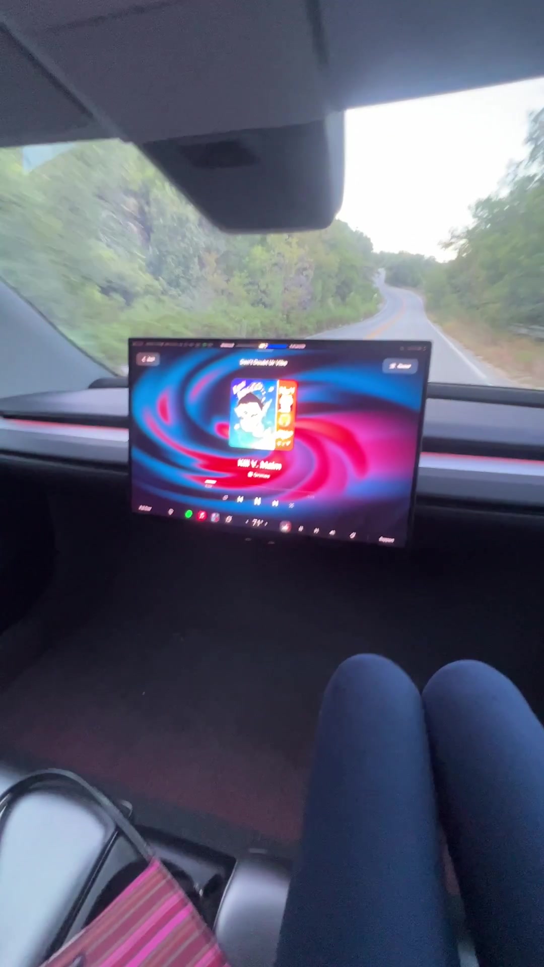 Passenger view of a wheel-less Cybercab cabin on a two-lane mountain road, center screen on, open footwell, striped bag on the seat.