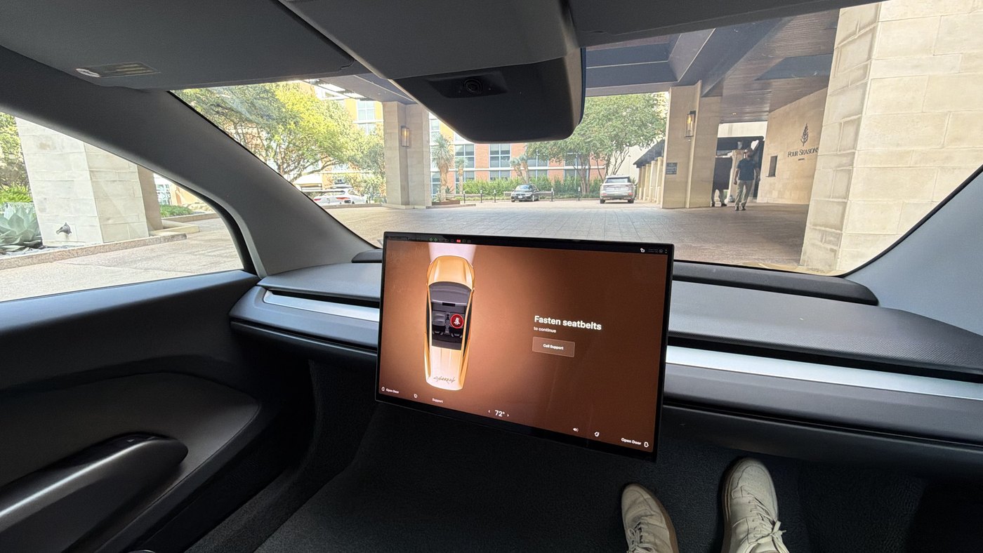 The inside of a Tesla Cybercab, with a centre touchscreen showing a gold Cybercab and a hotel entrance visible through the windscreen
