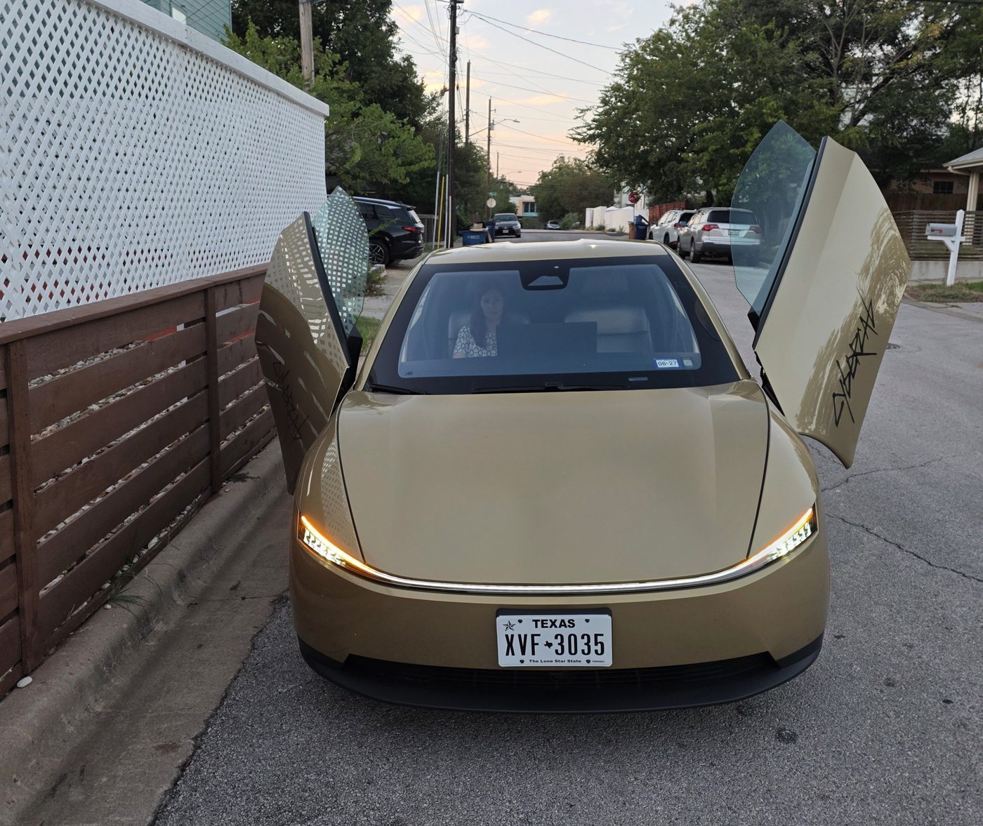 A silver Tesla Cybercab with its butterfly door partly open beside a fence on an Austin street