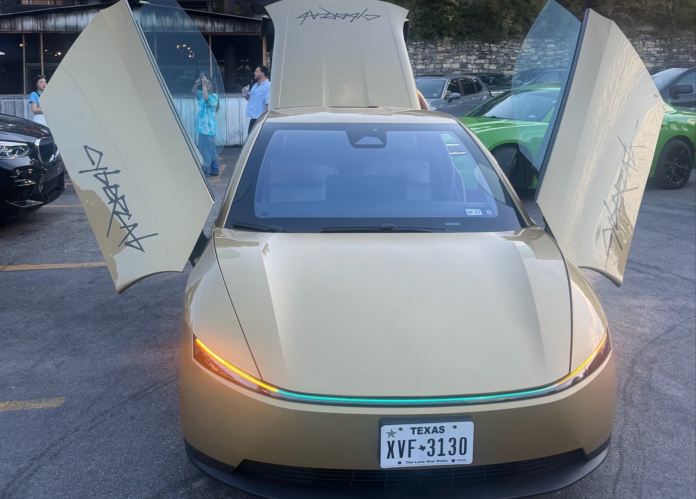A gold Tesla Cybercab with both doors and the rear hatch raised in a parking lot outside Terry Black's BBQ in Austin