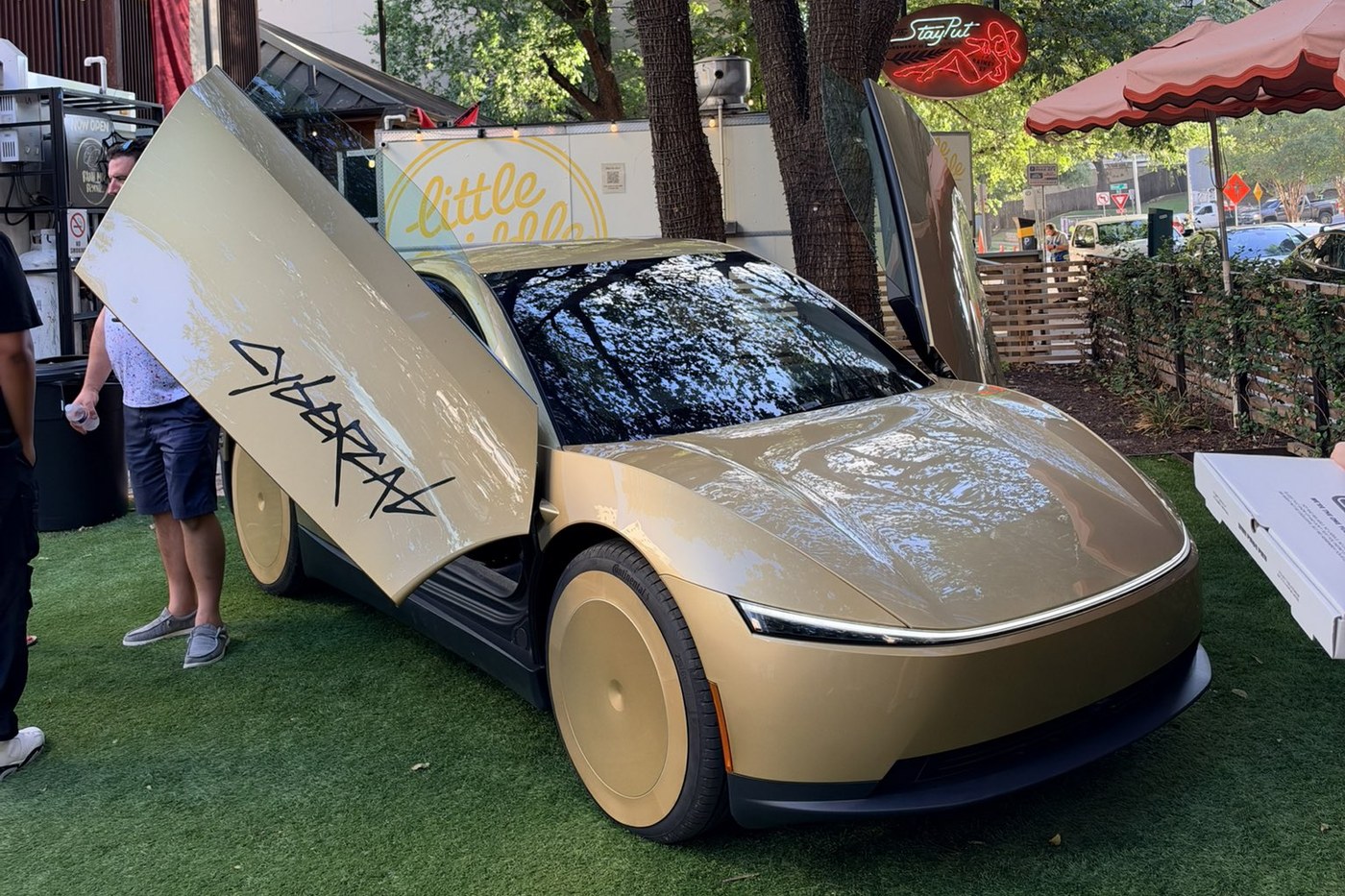 A gold Tesla Cybercab with its butterfly door raised, parked on artificial turf at a food truck park in Austin