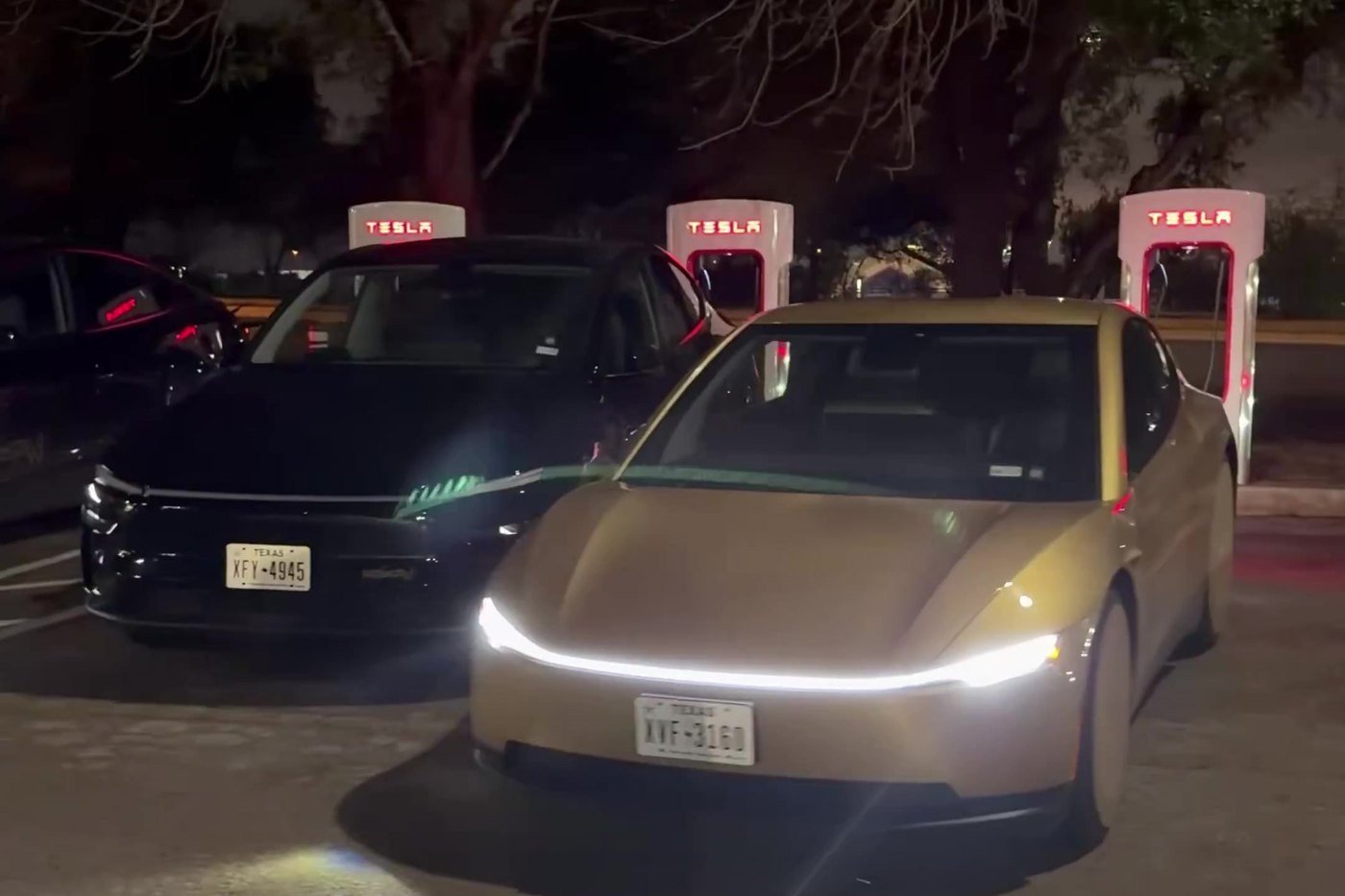 A gold Tesla Cybercab parked at a Supercharger stall beside a black Model Y robotaxi at night in Austin