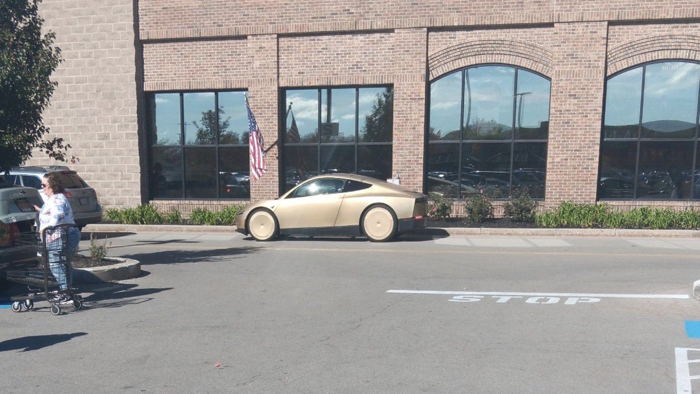 A gold Tesla Cybercab parked at the kerb outside a brick supermarket building with an American flag in Amherst, New York