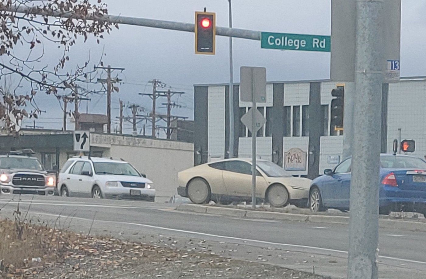 A gold Tesla Cybercab waiting at a red light beneath a College Rd street sign in Fairbanks, Alaska
