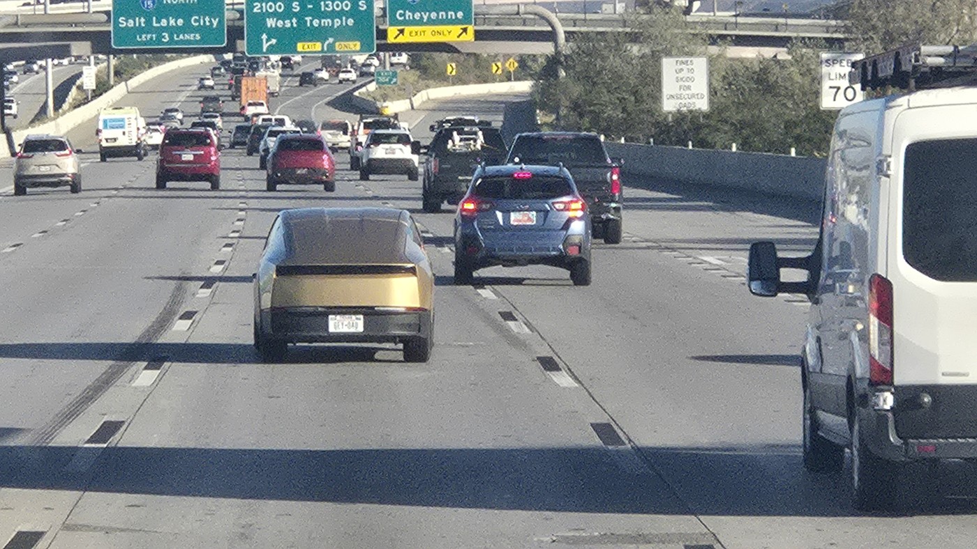 A gold Tesla Cybercab driving in interstate traffic in Salt Lake City beneath green overhead signs for Salt Lake City, West Temple and Cheyenne