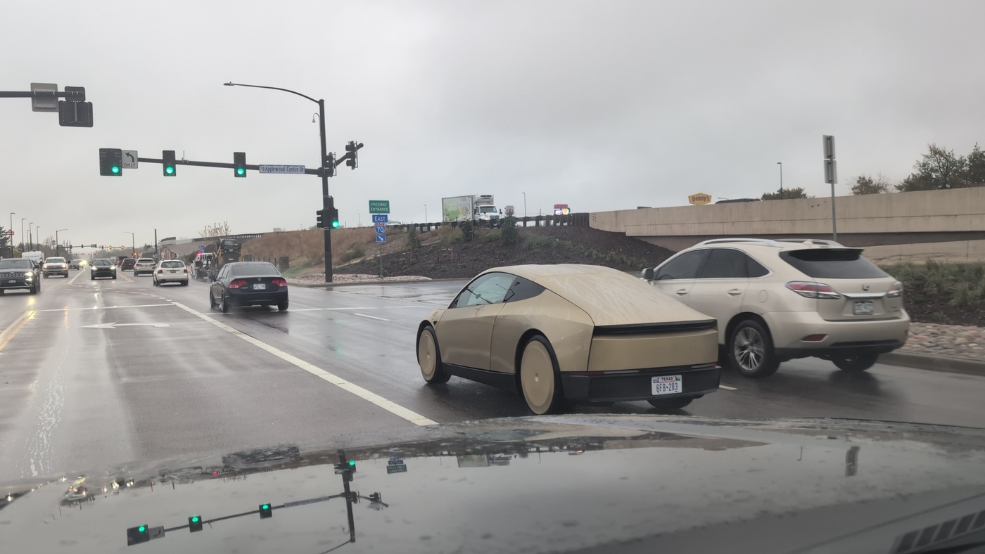 A gold Tesla Cybercab driving on a wet road in Wheat Ridge, Colorado, beside a white SUV under a gray sky