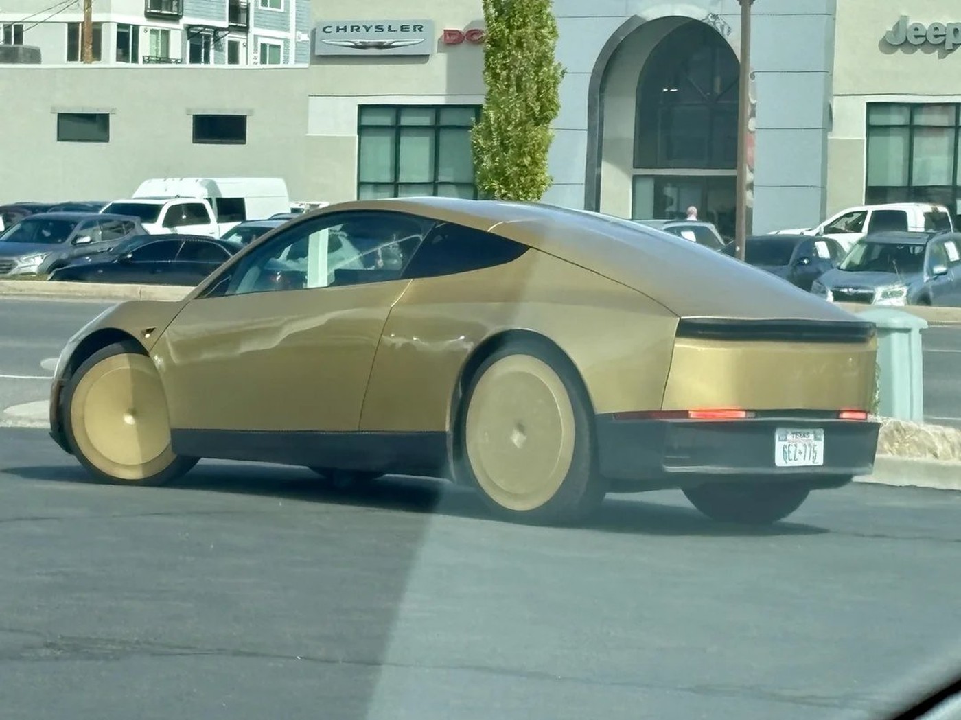 A gold Tesla Cybercab with Texas plates drives past a Chrysler and Jeep dealership in South Salt Lake, Utah
