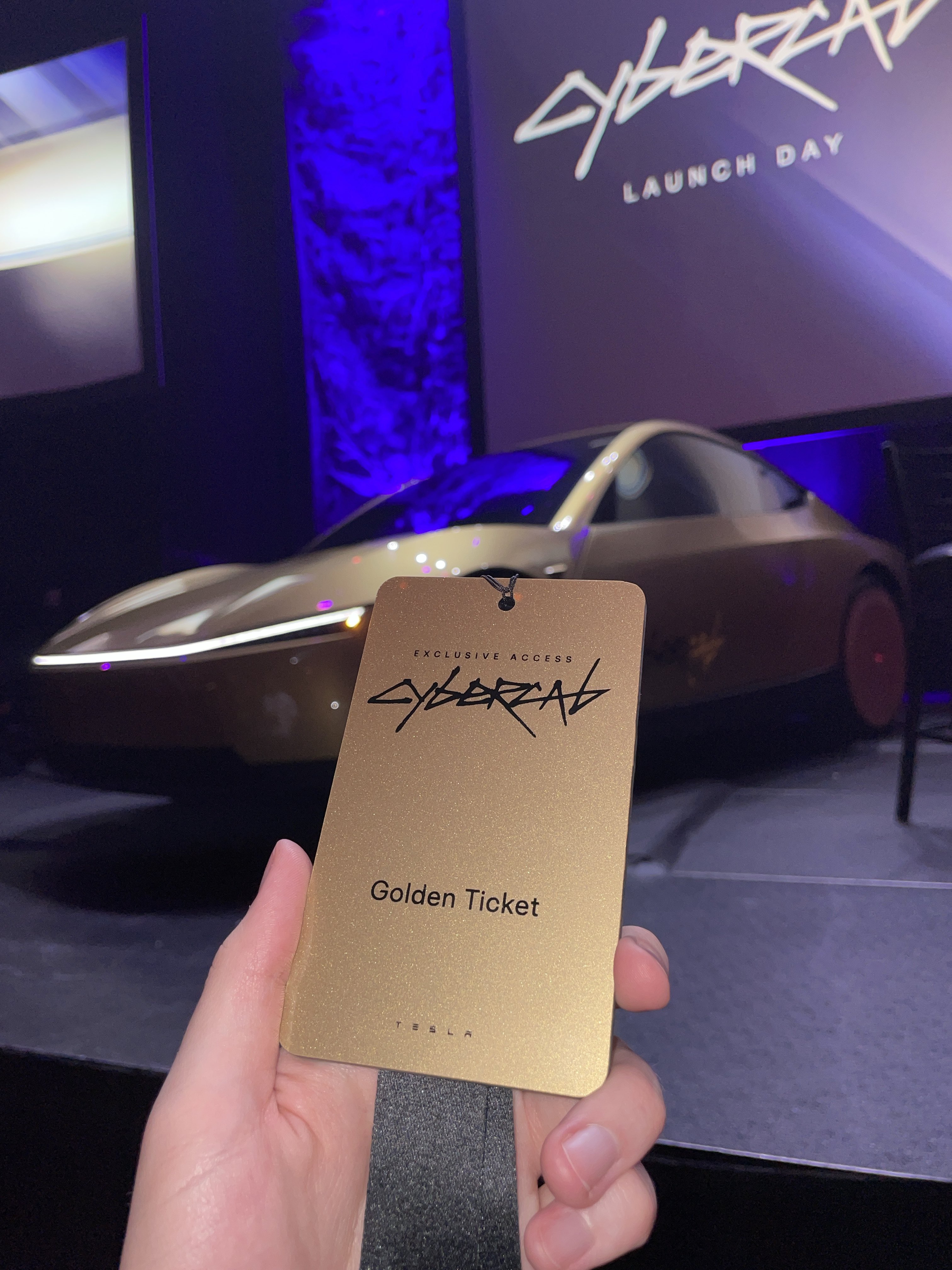 A gold Exclusive Access Golden Ticket badge held in front of a champagne Cybercab on the launch floor, CYBERCAB LAUNCH DAY on the screen behind