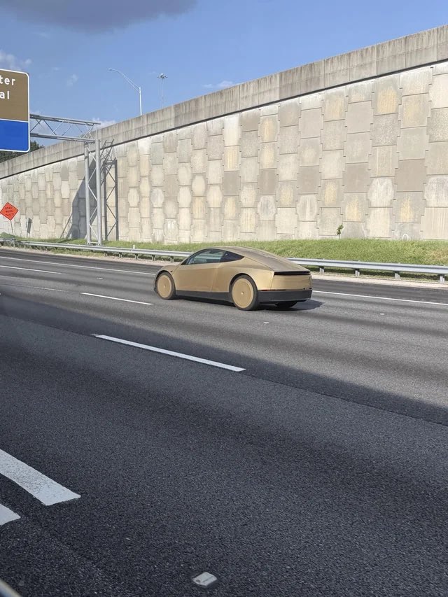 Gold Cybercab on a highway beside a sound wall.