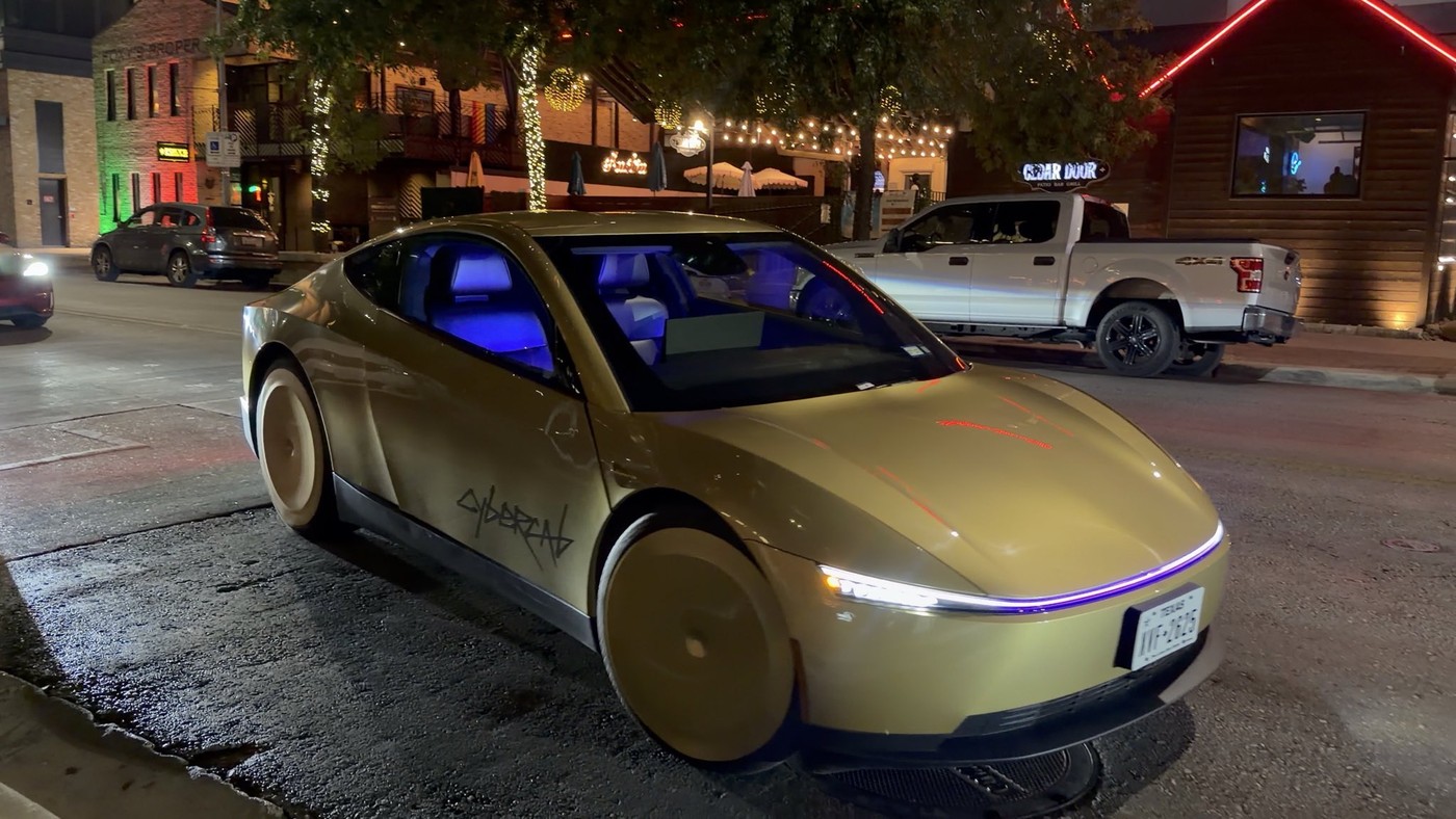 A gold Tesla Cybercab with its light bar glowing waits at the curb on an Austin street at night