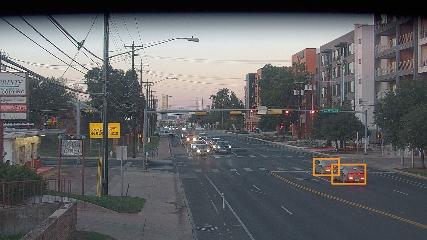 An Austin traffic camera frame at dusk with two Tesla Cybercabs highlighted by orange boxes as they cross an intersection