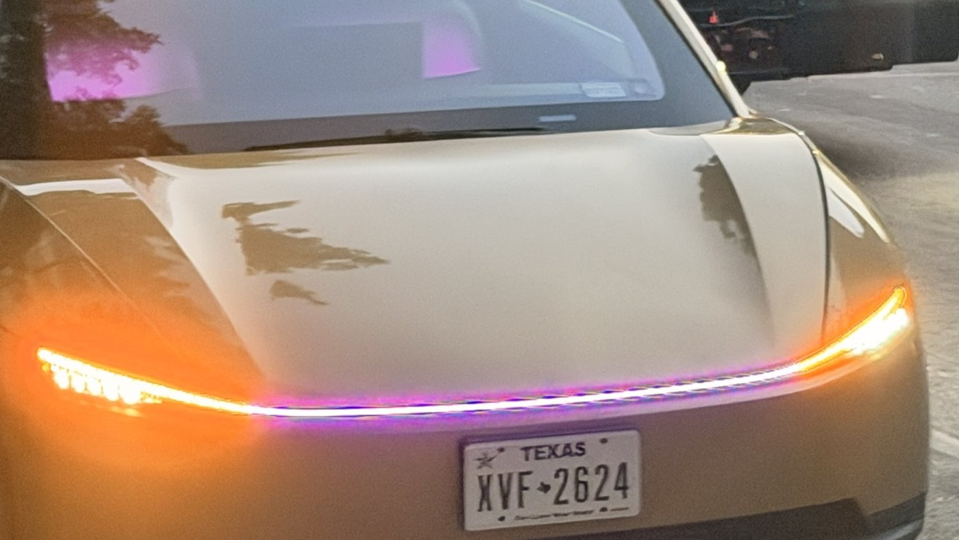 The front of a gold Tesla Cybercab with Texas plates, its full-width light bar glowing orange and purple