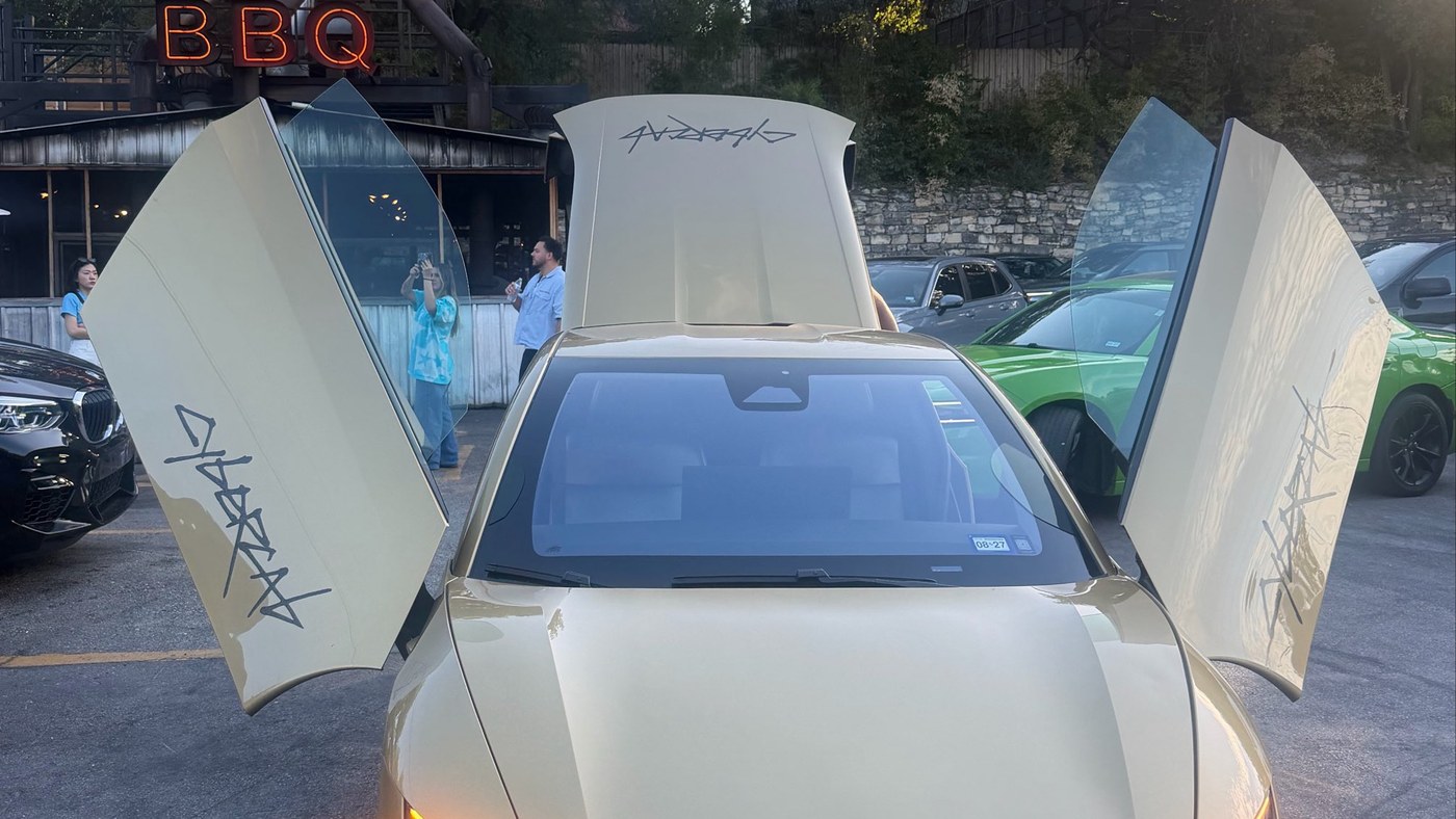 A gold Tesla Cybercab with butterfly doors and rear trunk open in an Austin parking lot