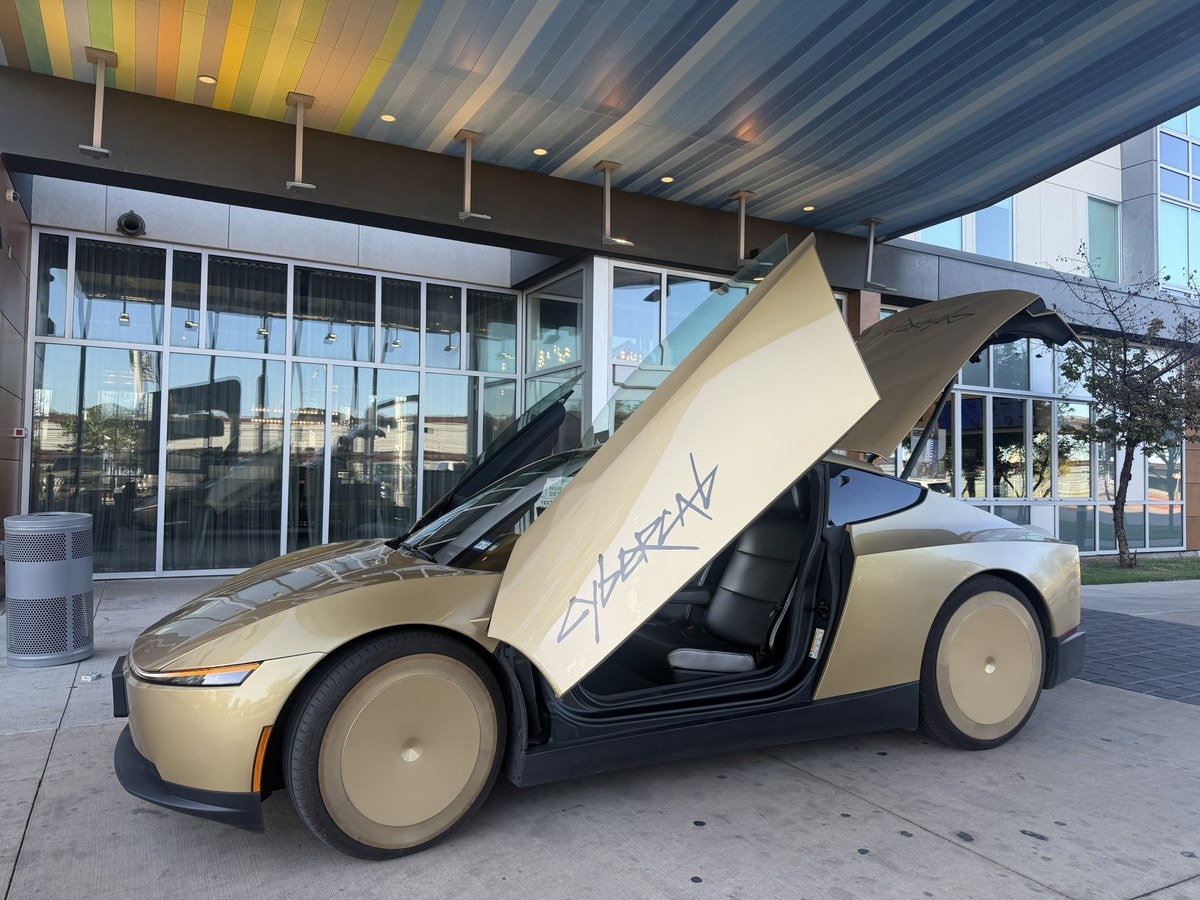 A gold Cybercab with butterfly doors and rear trunk raised at the curb, the hatch ready for bags. Photo via @BLKMDL3 / X