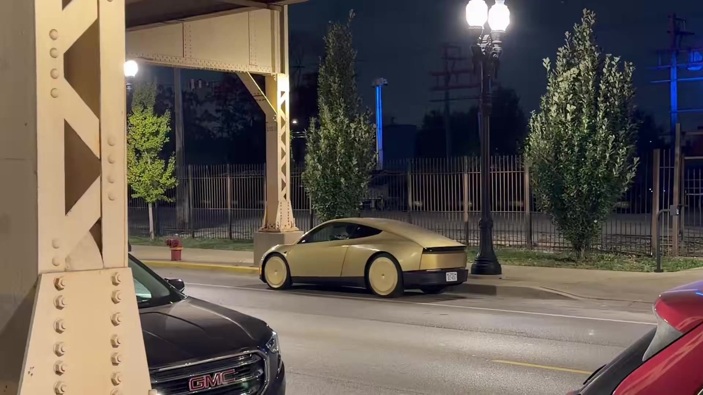 A gold Tesla Cybercab driving at night beside elevated rail tracks on Chicago's West Side