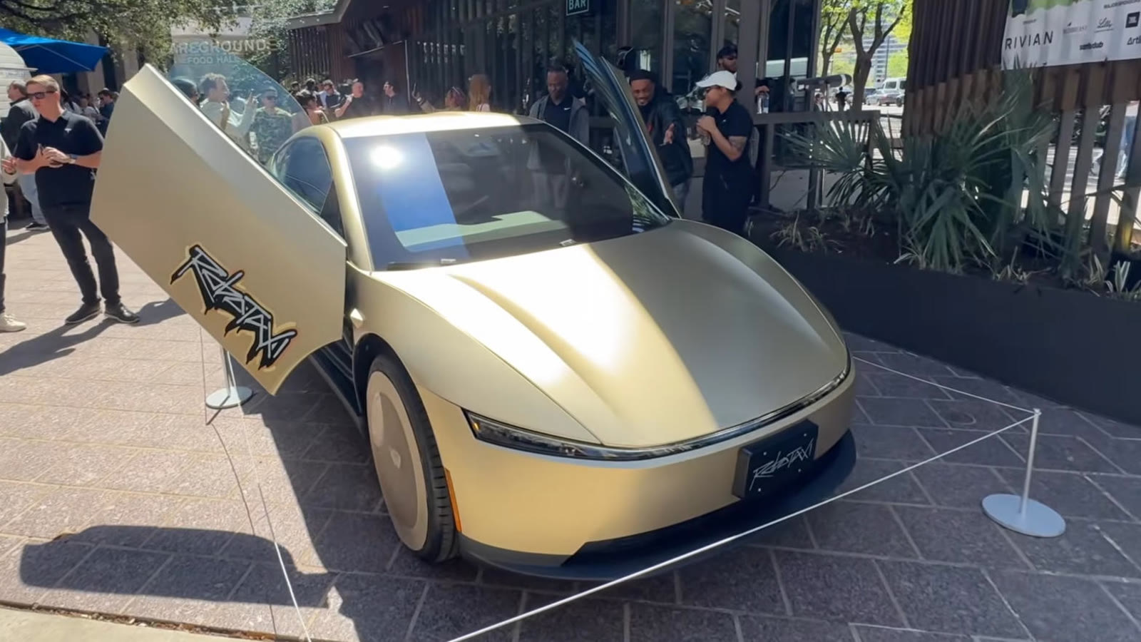 A Cybercab on display at an outdoor event with butterfly doors open and onlookers behind stanchions