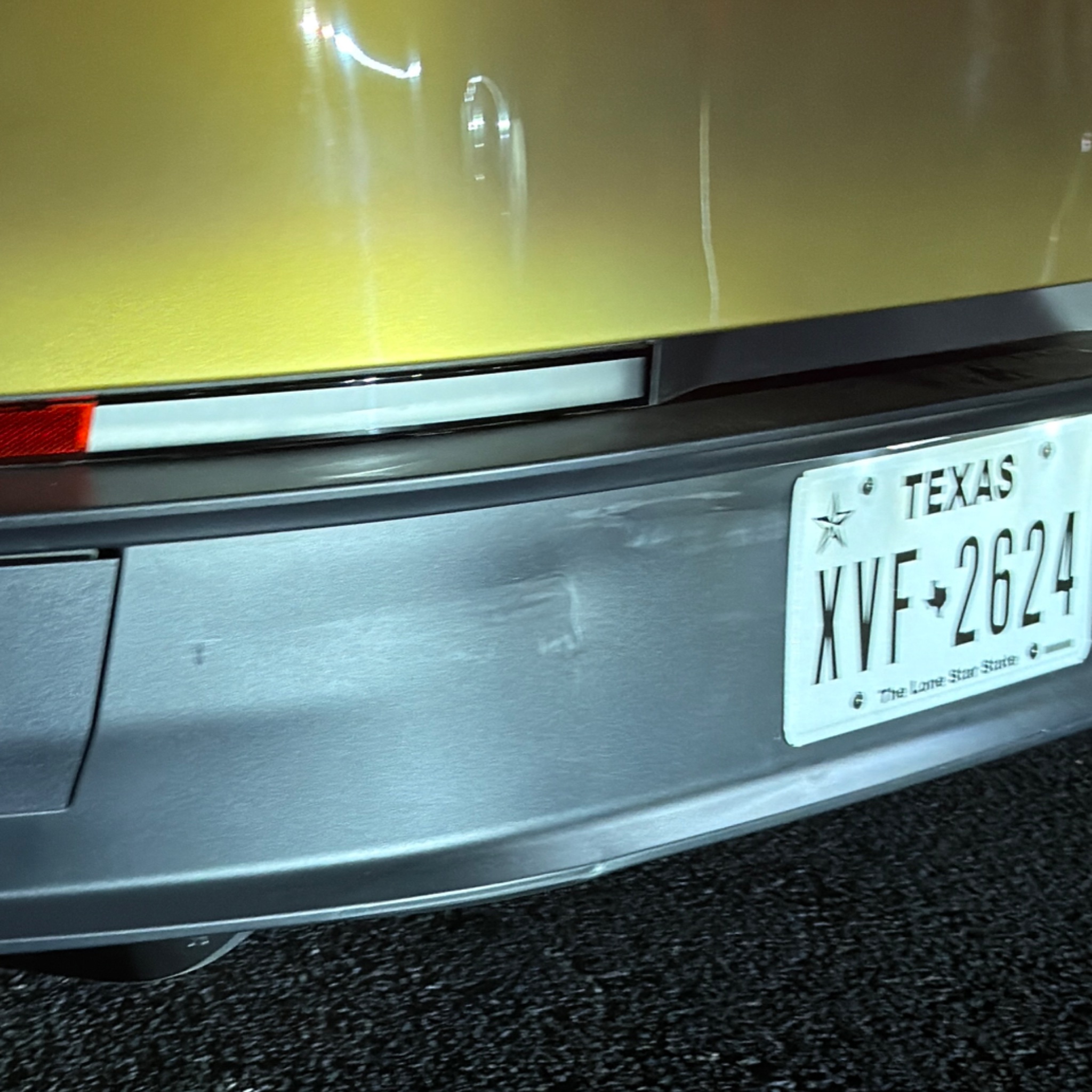 Close-up of a gold Cybercab rear bumper with a light scuff and small dent left of Texas plate XVF-2624