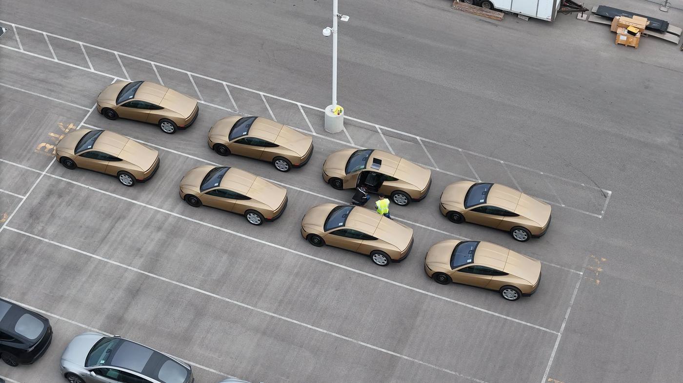 Nine gold Cybercabs staged in a Giga Texas lot, one with a butterfly door open and staff alongside.