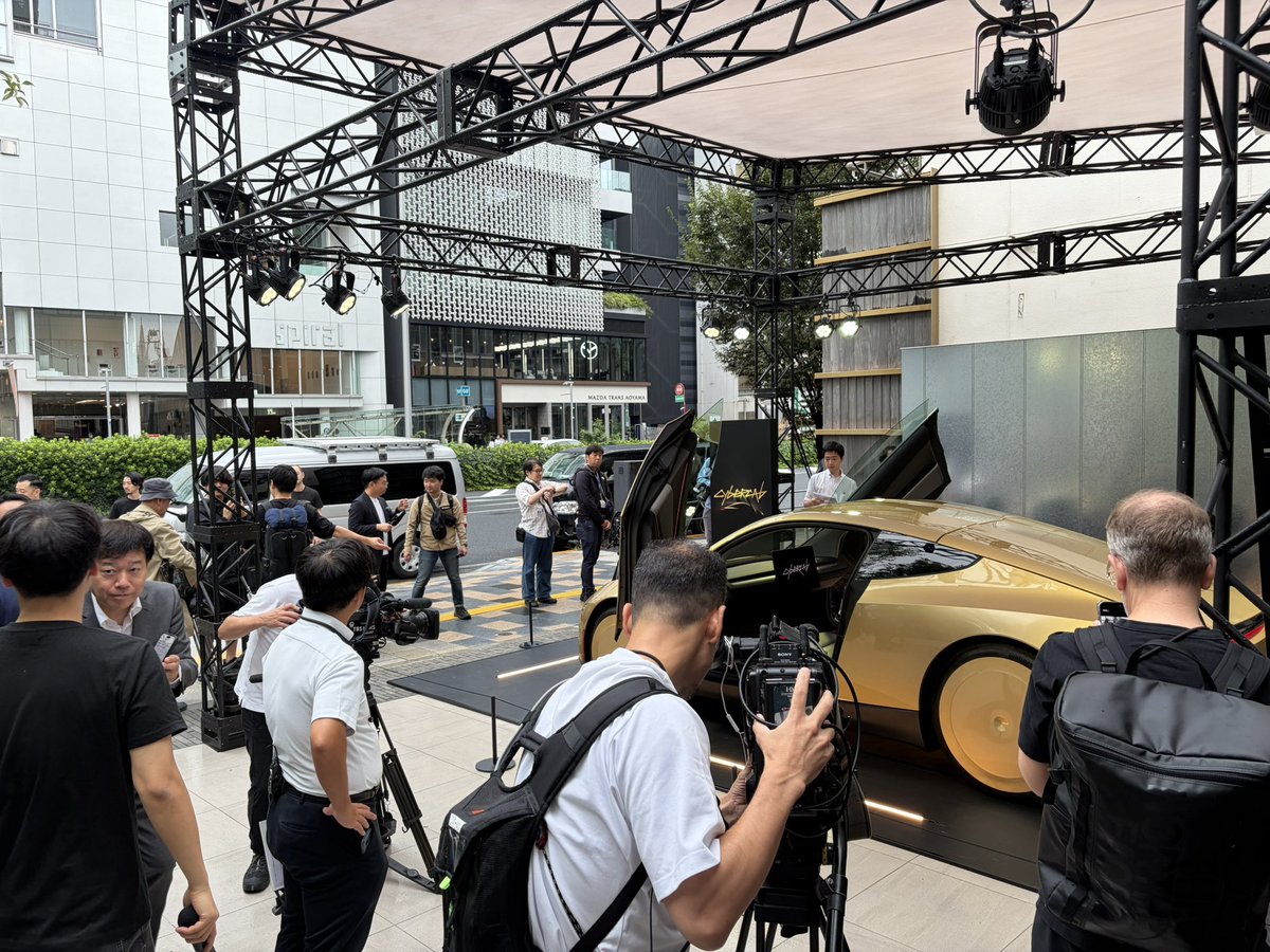 A gold Cybercab on a wooden deck in Aoyama, butterfly doors closed, with visitors and cameras around the car.