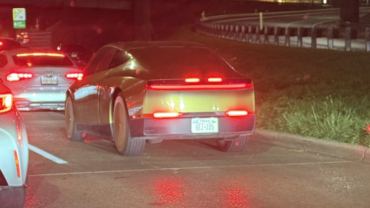 A gold Tesla Cybercab in evening traffic on a Texas road, seen from behind with its full-width rear light bar lit