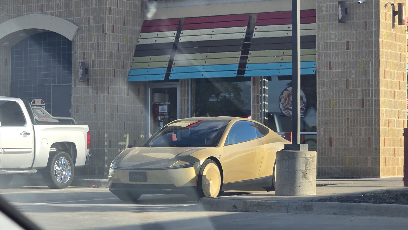 A gold Tesla Cybercab parked outside a street taco restaurant beside a white pickup truck at the Westmoreland Supercharger in Dallas