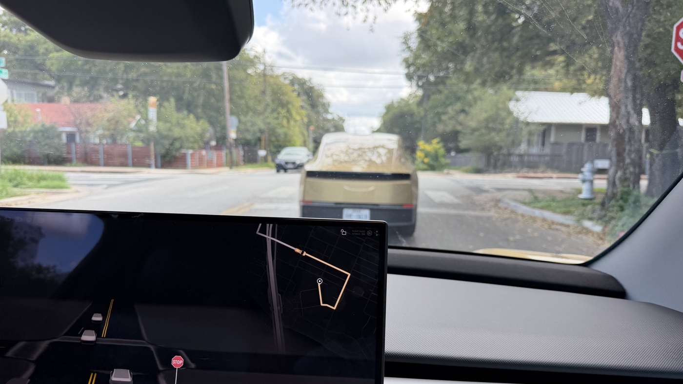 The view from inside a Tesla Cybercab, with its center screen showing the route and another gold Cybercab driving directly ahead on a leafy street