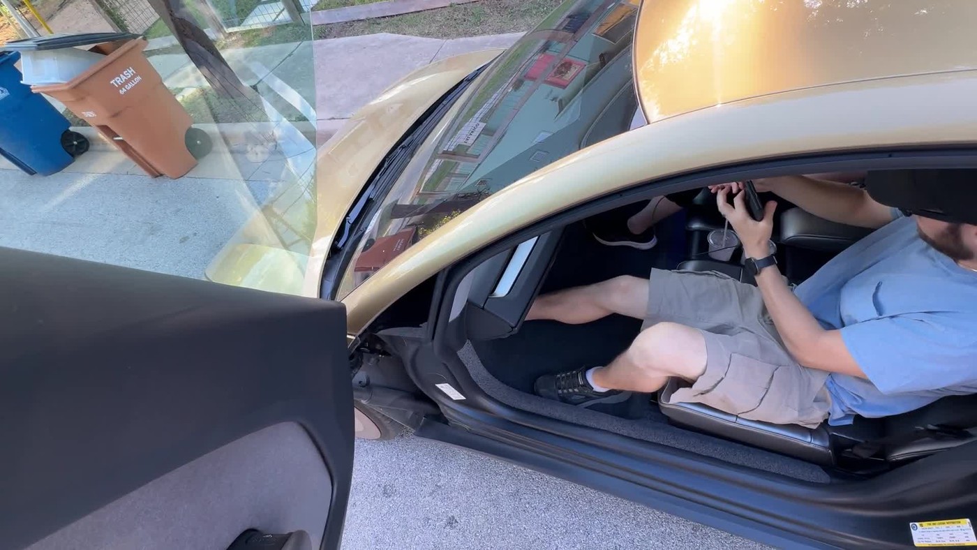 A gold Tesla Cybercab with its butterfly door open and a rider seated inside, parked at a curb