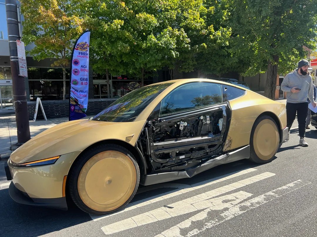 A gold Tesla Cybercab parked on a Philadelphia street with the outer skin of its driver door missing, exposing the door frame and wiring