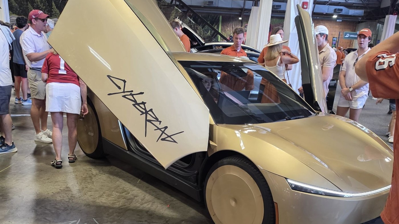 A gold Tesla Cybercab with both butterfly doors open, on display indoors at the State Fair of Texas in Dallas