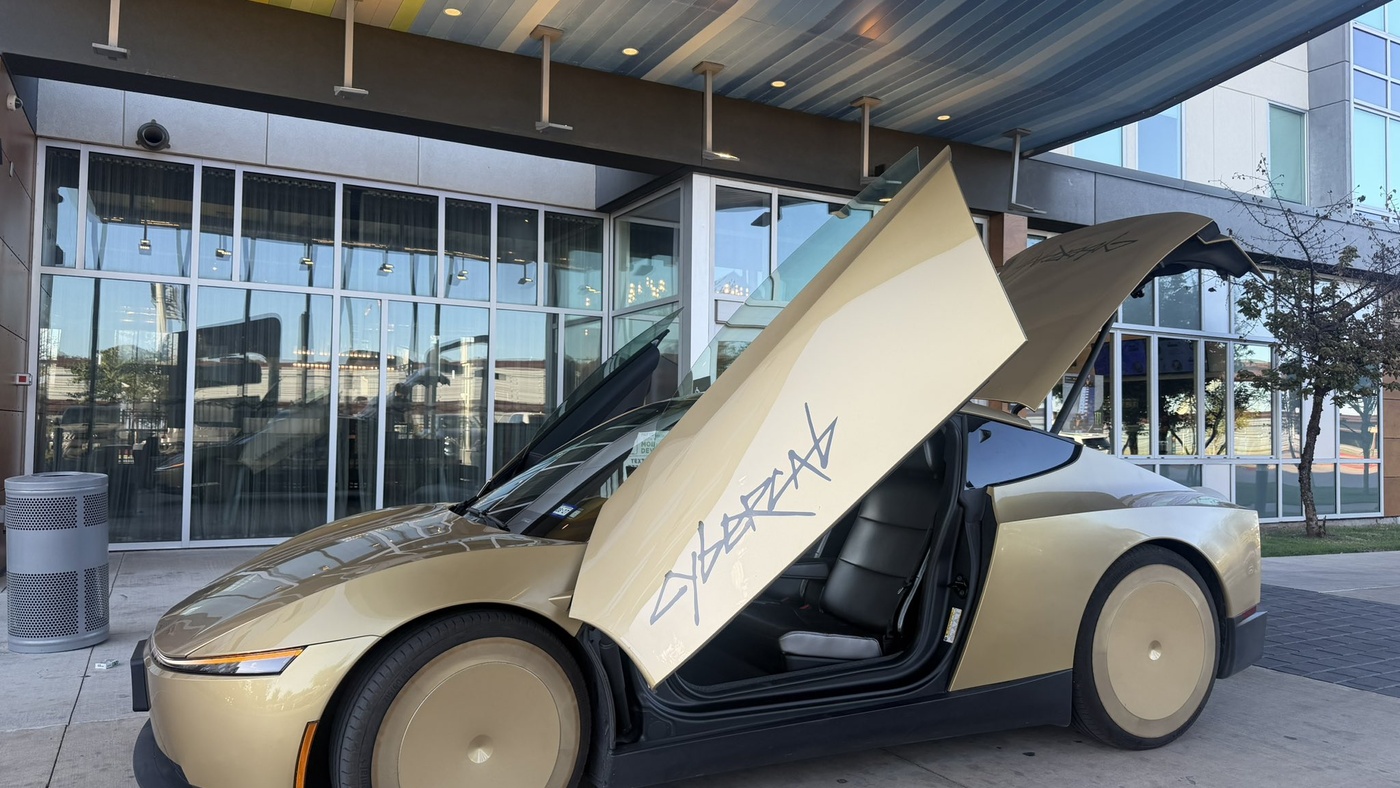 A gold Tesla Cybercab parked under a building overhang with butterfly doors and rear trunk hatch open