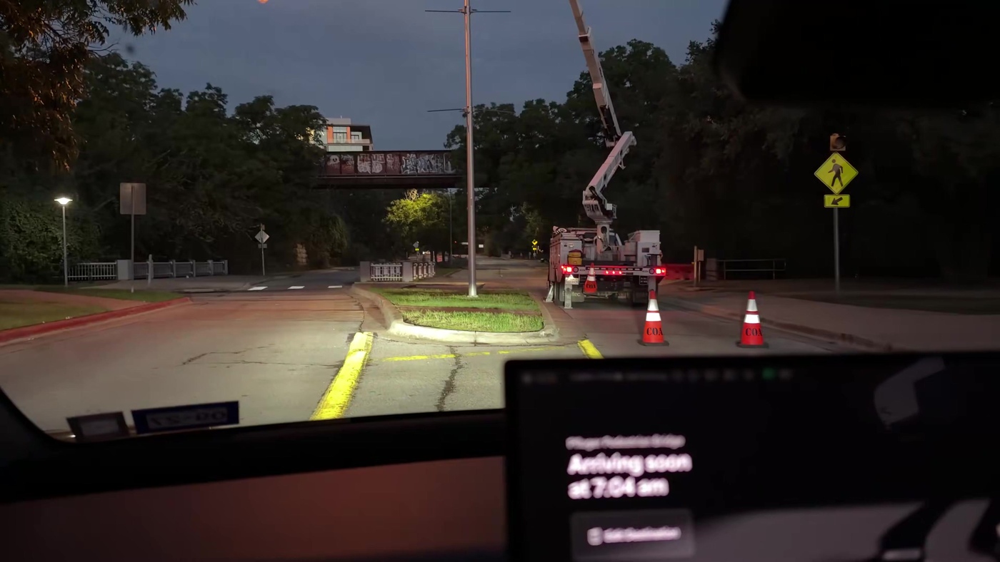 View from inside a Cybercab at dawn as it approaches a utility bucket truck and orange cones, with the ride screen reading Arriving soon at 7:04 am