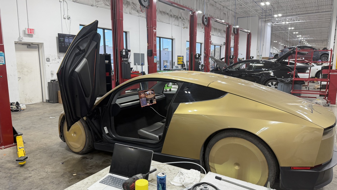 A gold Tesla Cybercab with its butterfly door raised in a Tesla service bay, with a cable running from its cabin to a workbench holding a laptop and tools