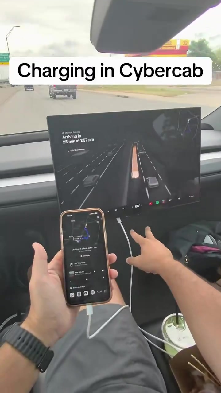 A passenger in a wheel-less Cybercab holds a charging iPhone on a white USB cable plugged in under the center display while the coupe drives on a highway.